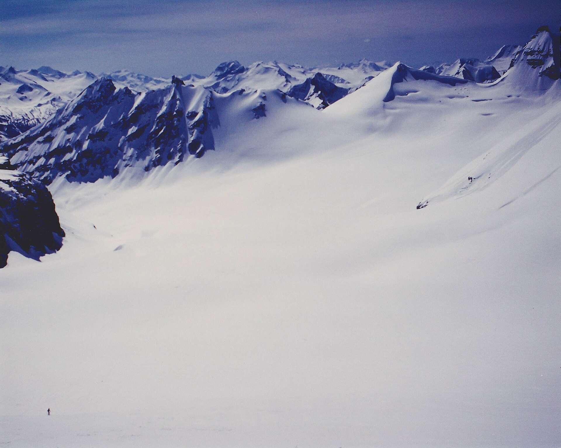 A lone skier crossing a vast glaciated basin below jagged peaks in the Selkirk Mountains
