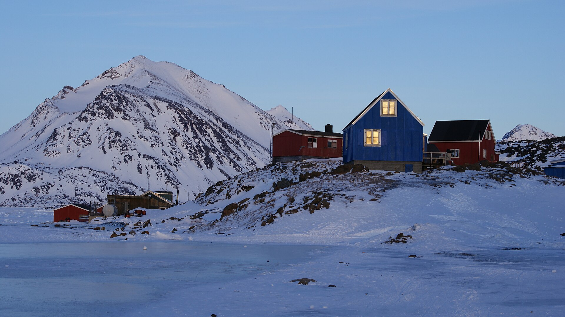 A blue house beneath snow-covered mountains at winter sunset in Kulusuk