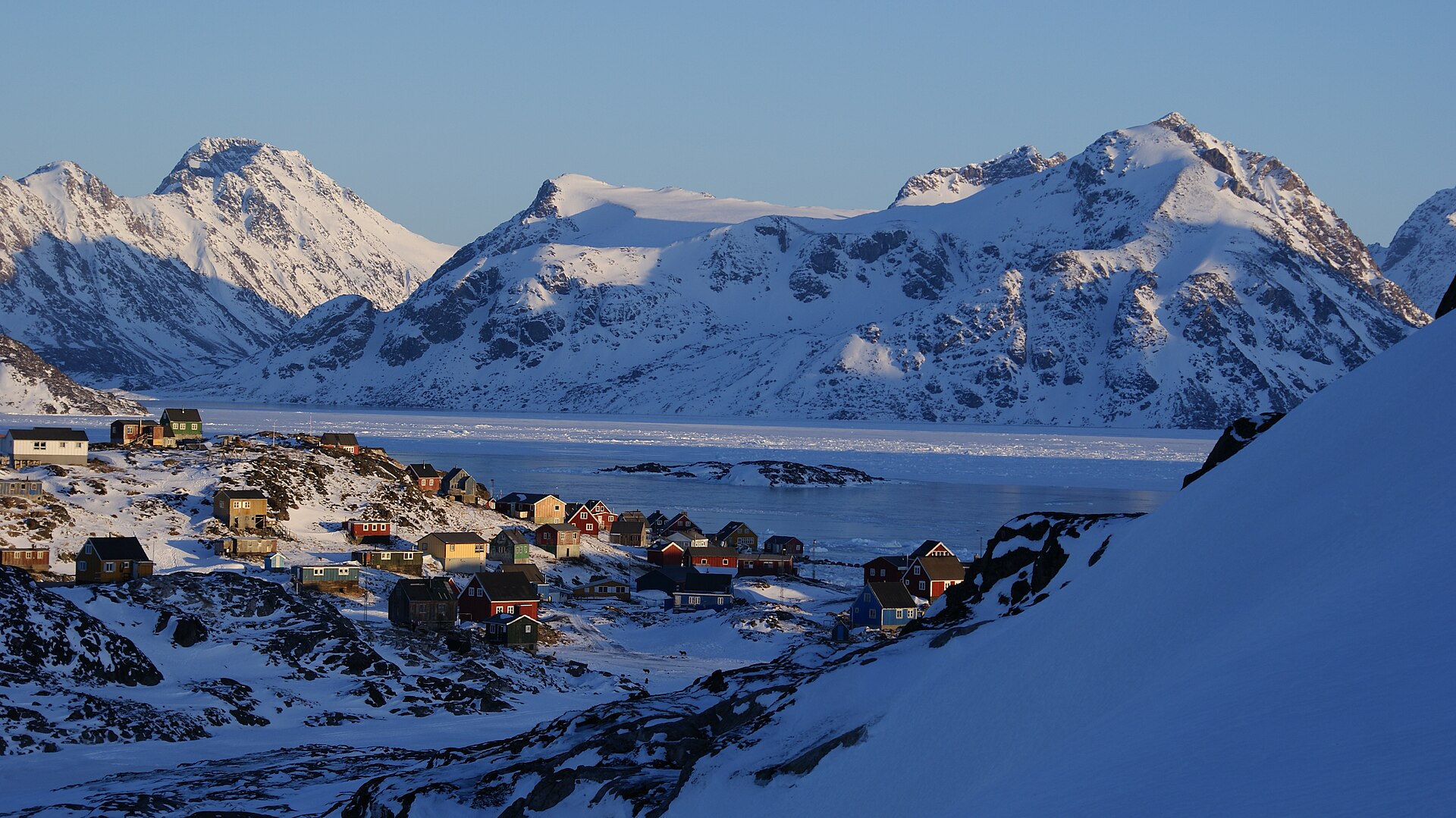 Kulusuk settlement surrounded by snow with mountainous Apusiaajik Island behind