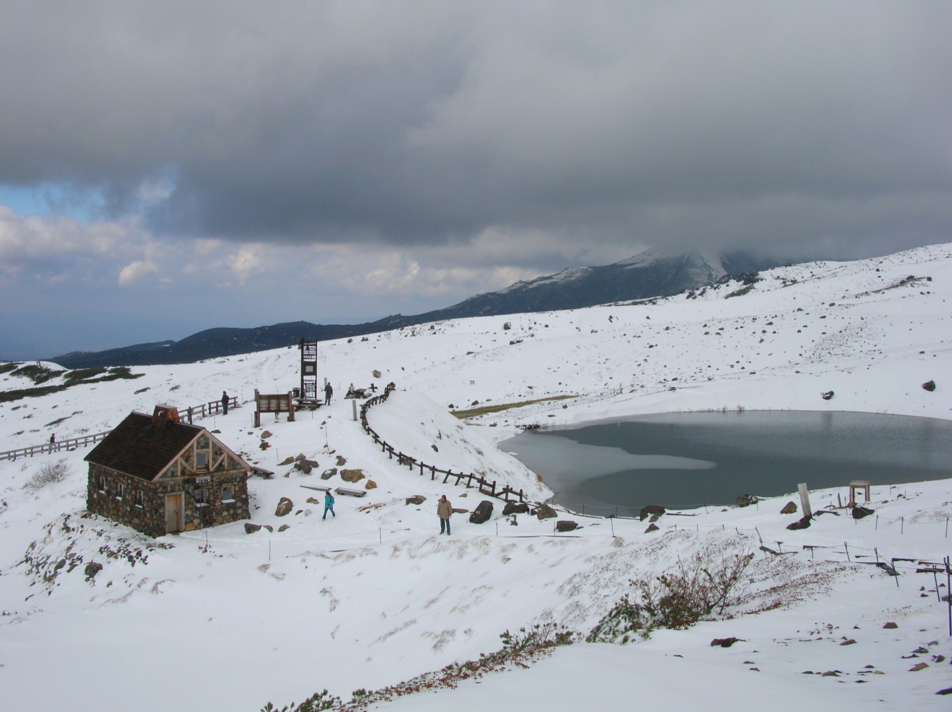 A stone hut and small pond on a snow-covered Daisetsuzan plateau, with a cloud-capped peak behind