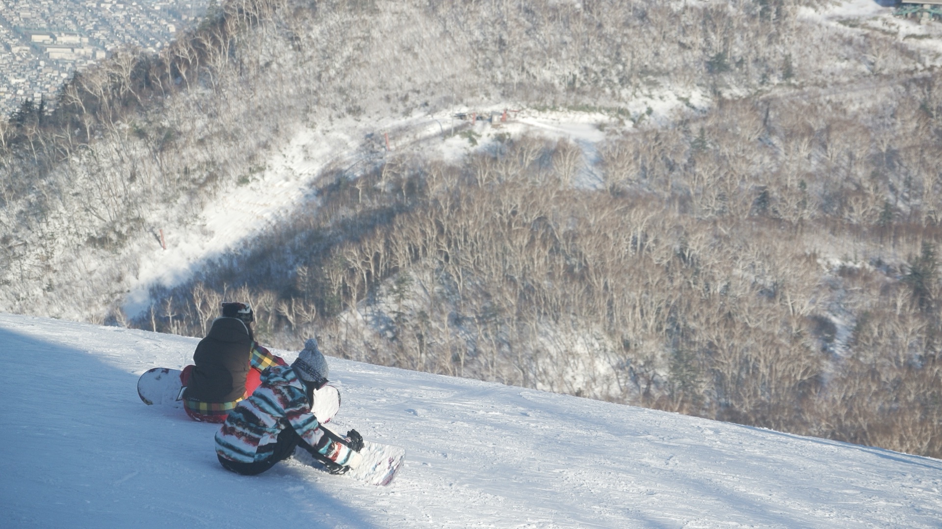 Two snowboarders resting on a pisted slope at Sapporo Teine, high above the city below
